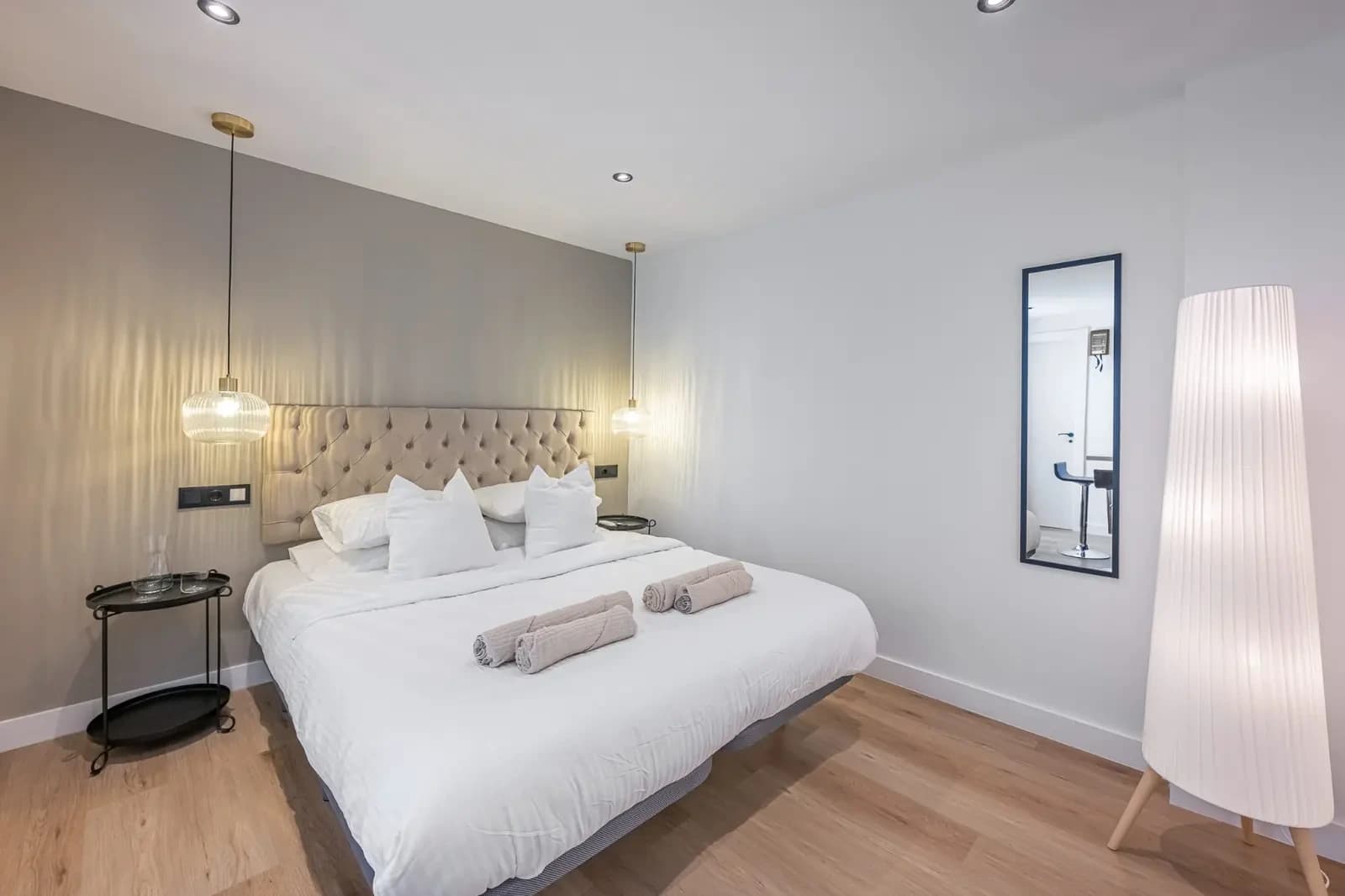 A suite bed with a tufted linen headboard between two hanging pendant lights, a floor lamp and a tall mirror alongside