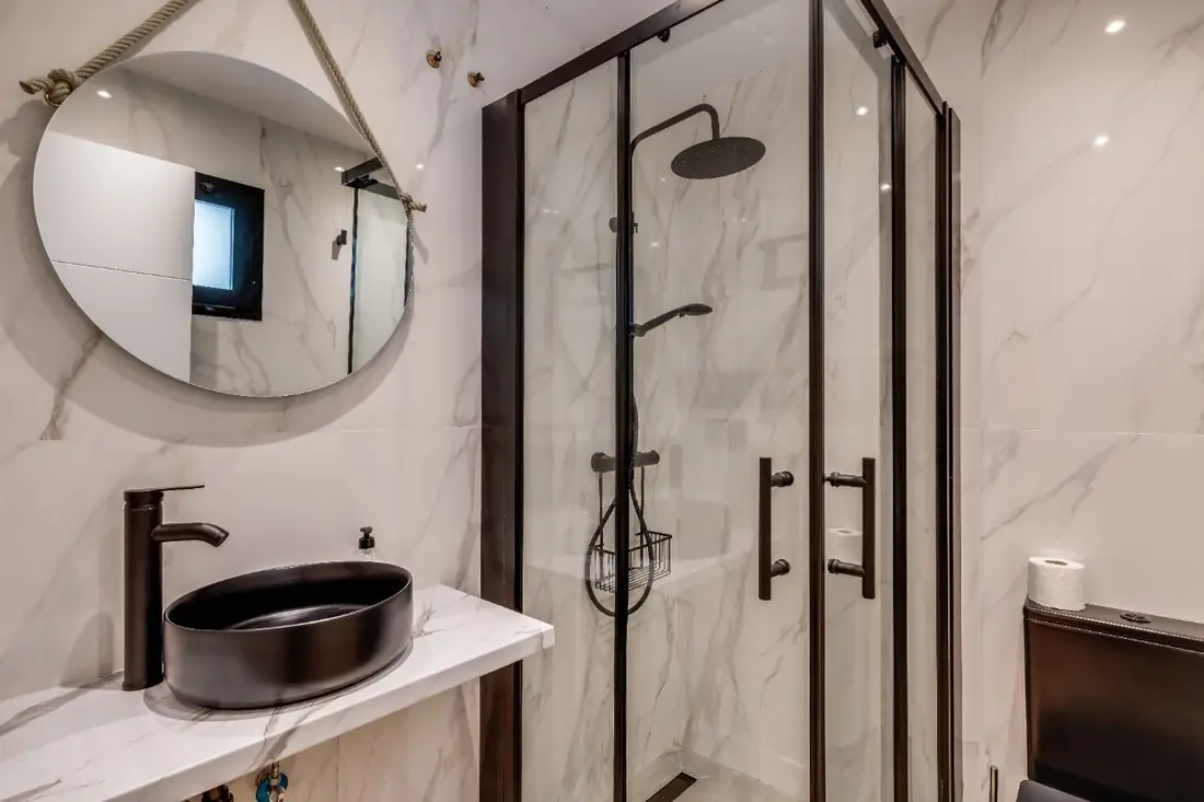 The Micro Suite bathroom: a round mirror above a black stone basin on marble, beside a glass walk-in shower