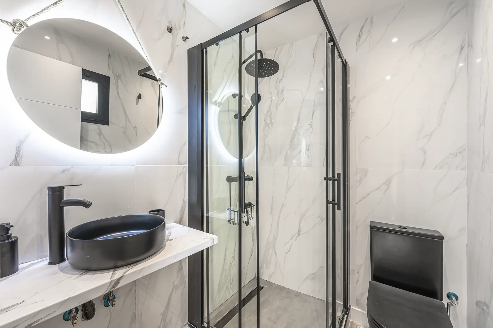 Room 2: a round lit mirror and black basin on marble, beside a corner shower in black framed glass