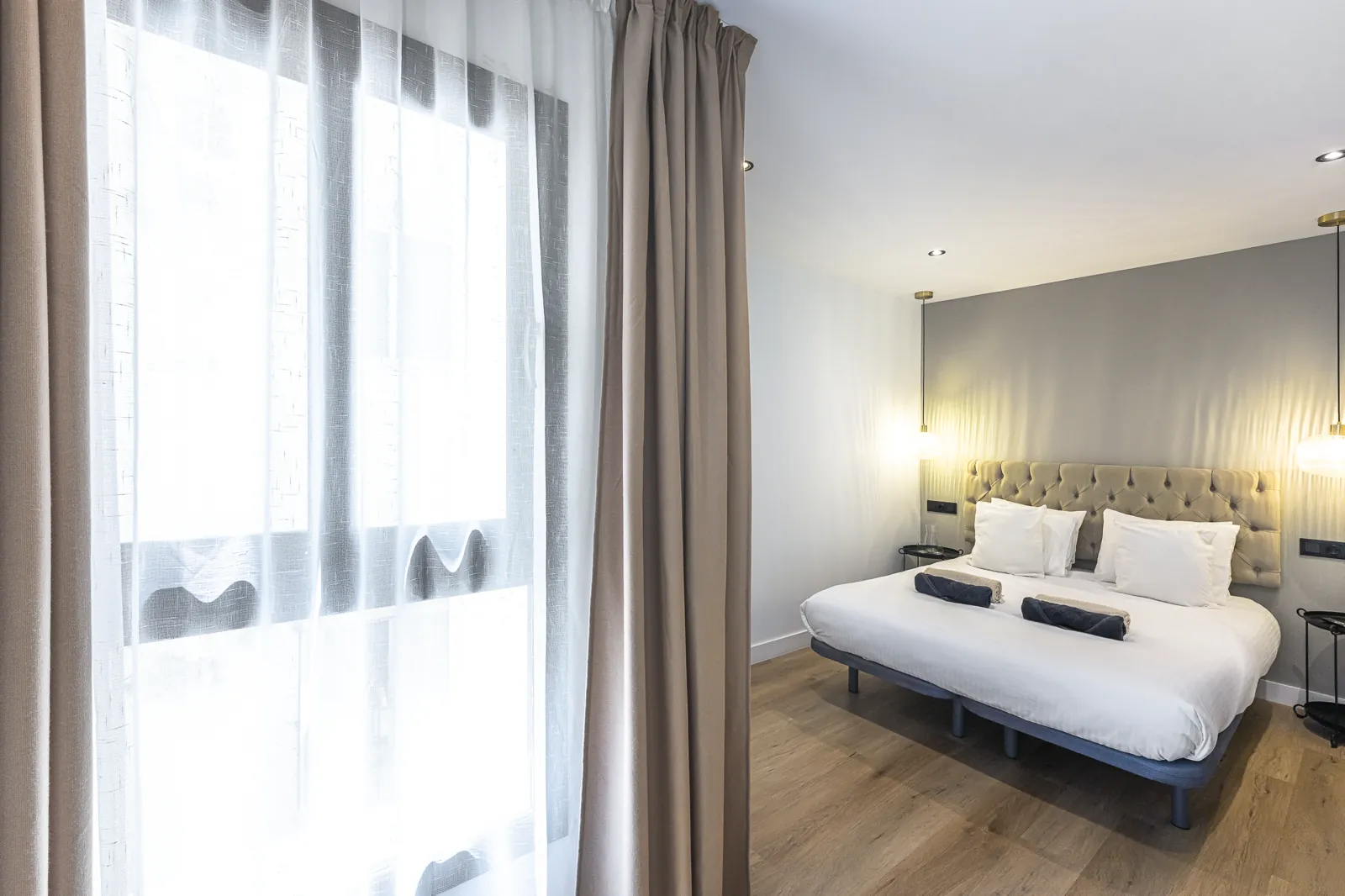 The Signature Suite: the bed beside a tall window in linen curtains, with reading lights on the headboard wall
