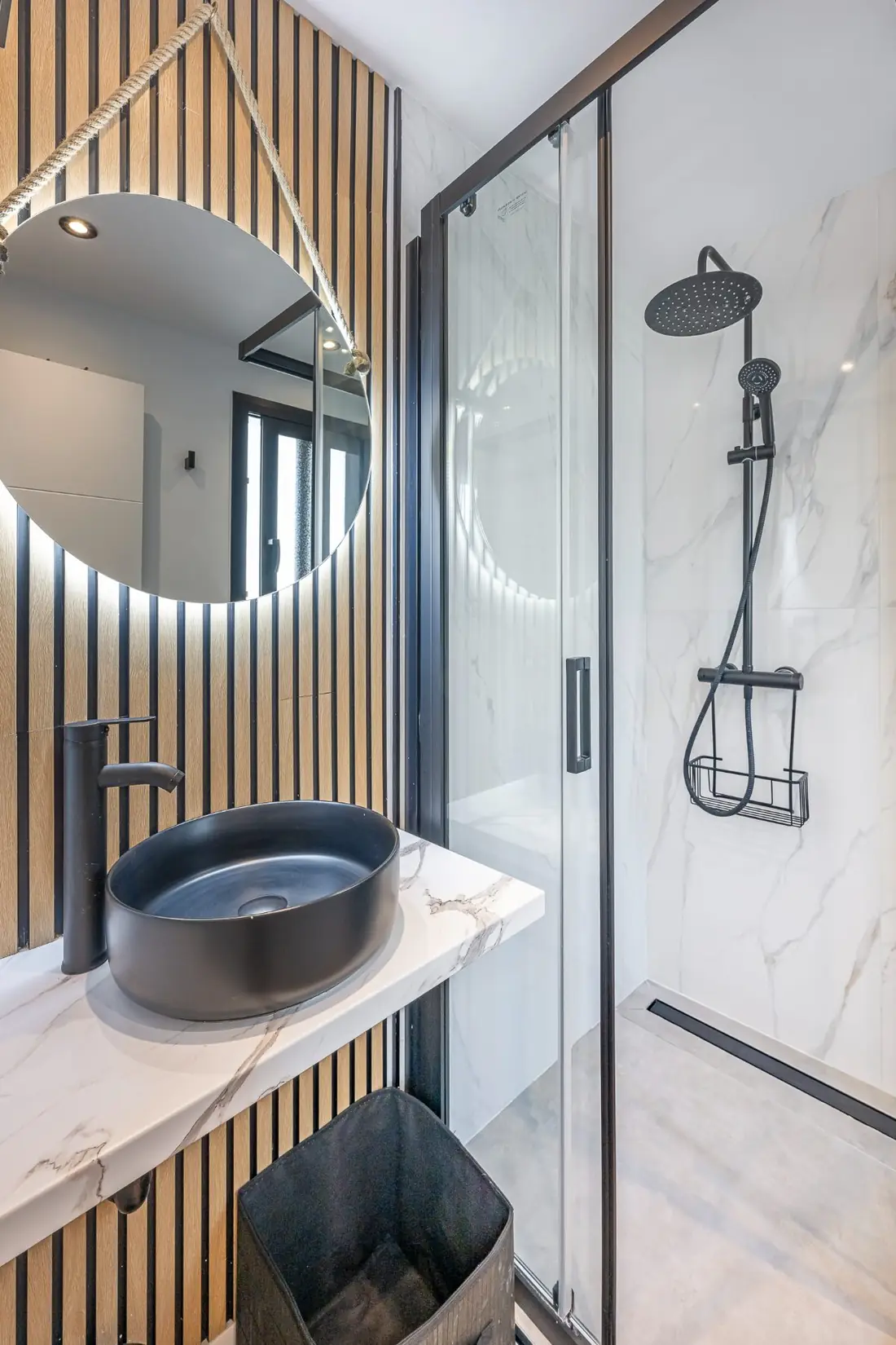 The bathroom: a round lit mirror and black stone basin against oak slats, next to the glass shower