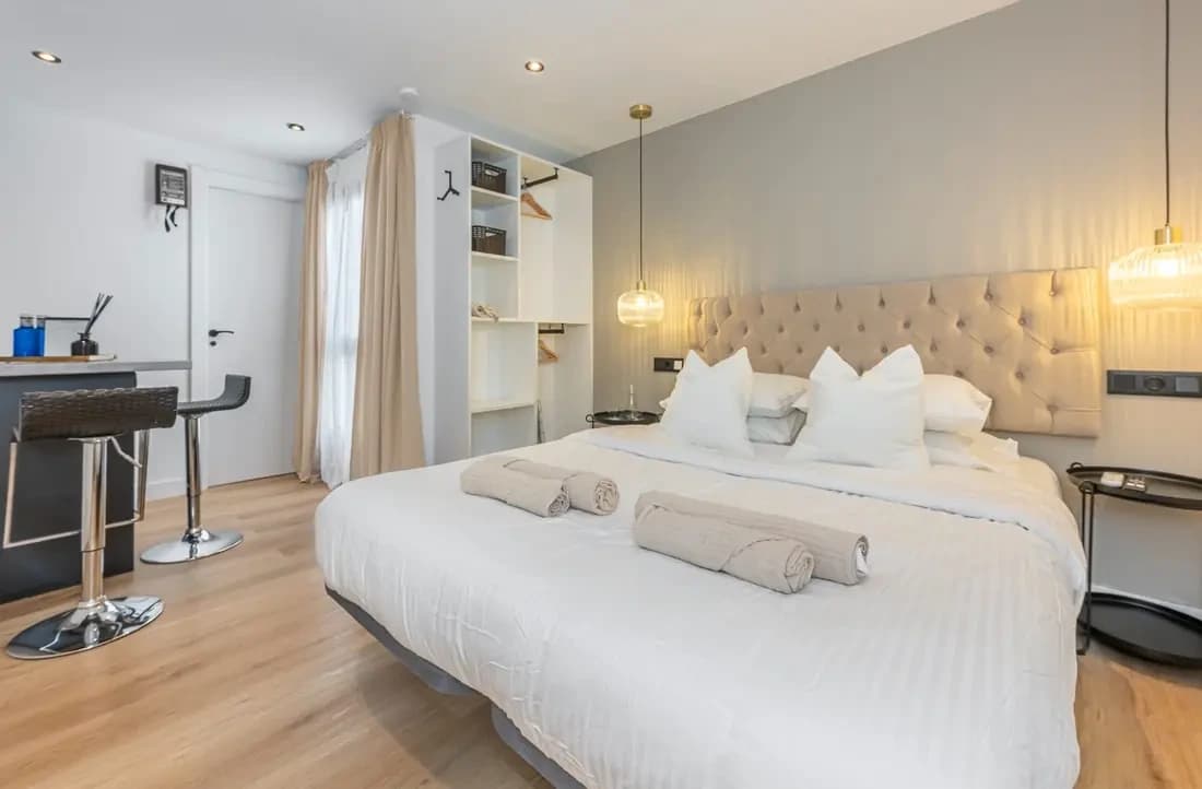 The Signature Suite: a bed with a tufted headboard, pendant lights, open shelving and a breakfast bar with two stools by the curtained window